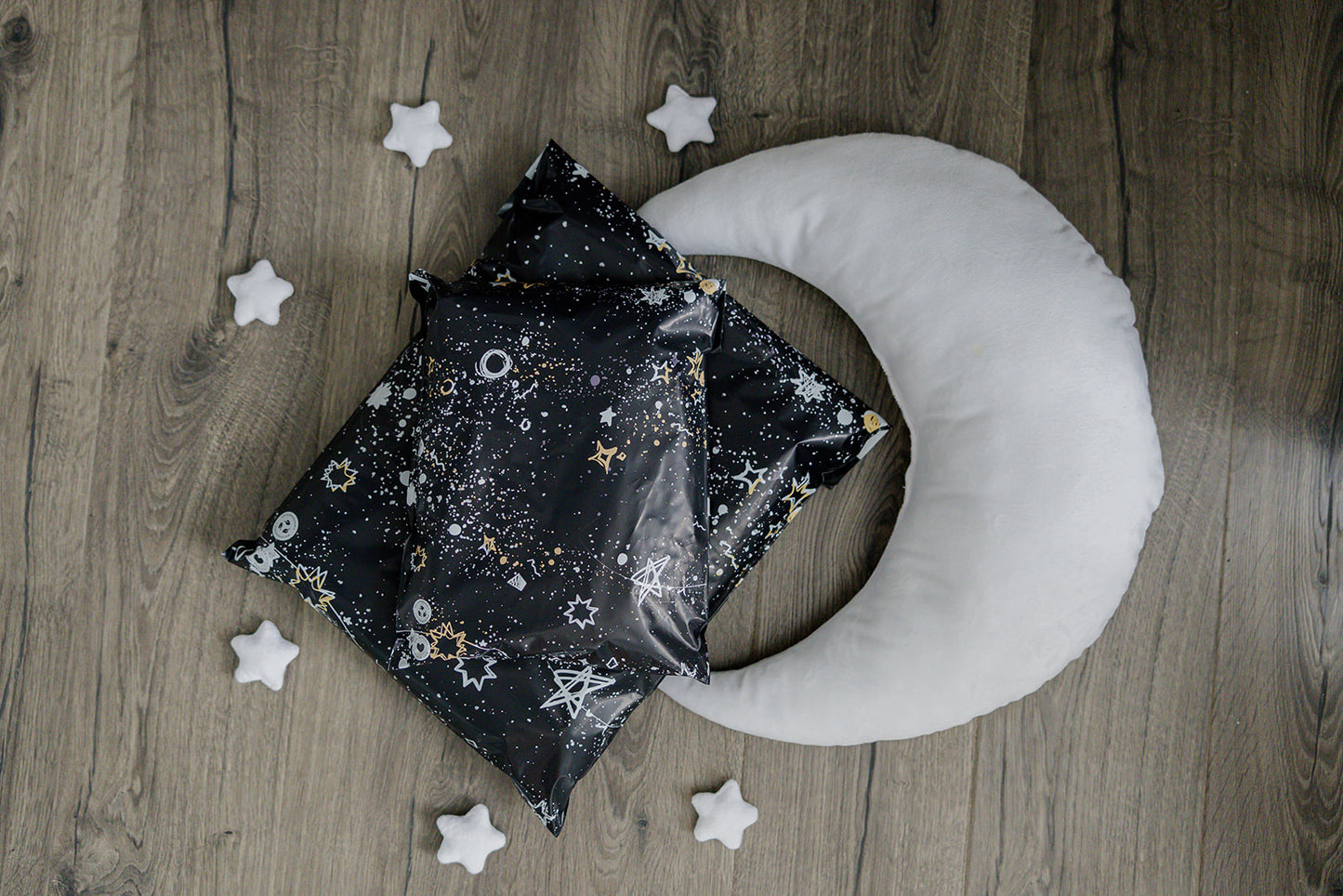 A recyclable Midnight Galaxy Mailers 10" x 13" pillow with stars and a crescent design laid on a wooden floor by impack.co.