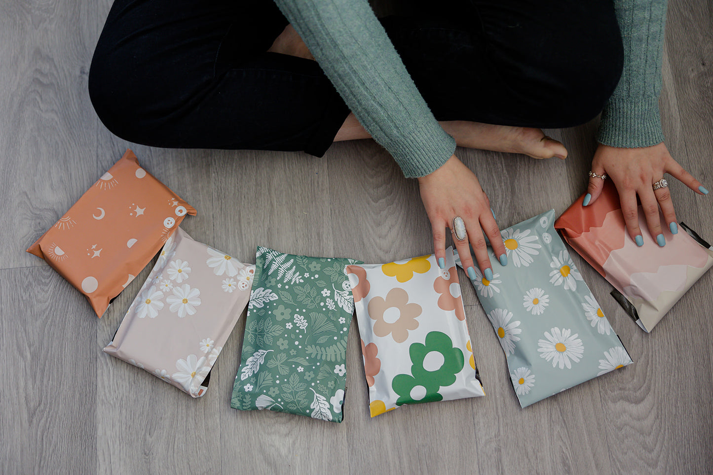 A woman is sitting on a wooden floor with a bunch of impack.co Biodegradable Mailers 10" x 13" of different colors.