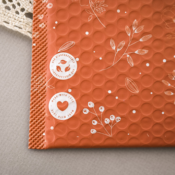 A bubble mailer with a nature-inspired design in orange, featuring printed symbols and text: "100% biodegradable" with a leaf icon, and "Made with love" with a heart icon.