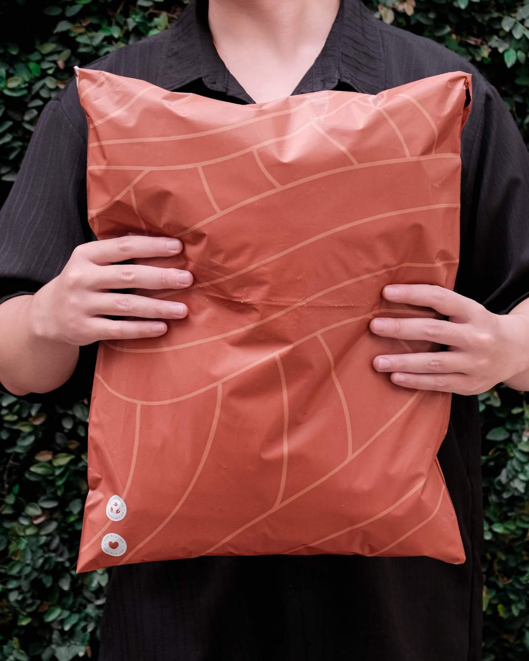 A person in a dark shirt holds a large, orange, patterned package with both hands in front of green foliage.
