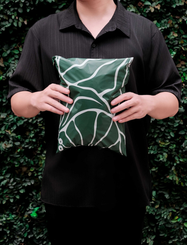 A person in a black shirt holds a green and white patterned pillow against a leafy background.