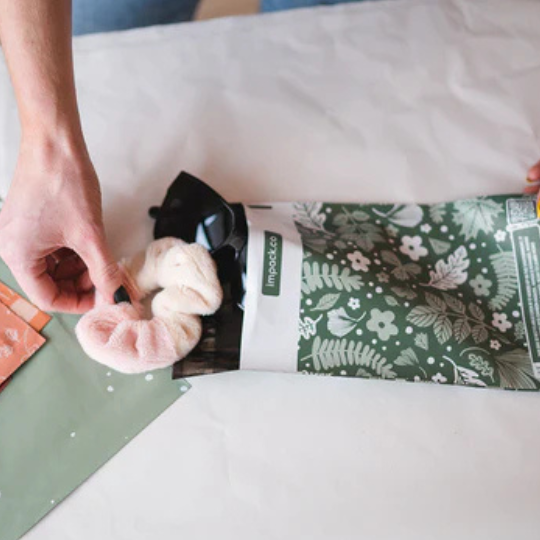 A person places a soft, pink scrunchie into a green floral-patterned mailing bag on a white surface.