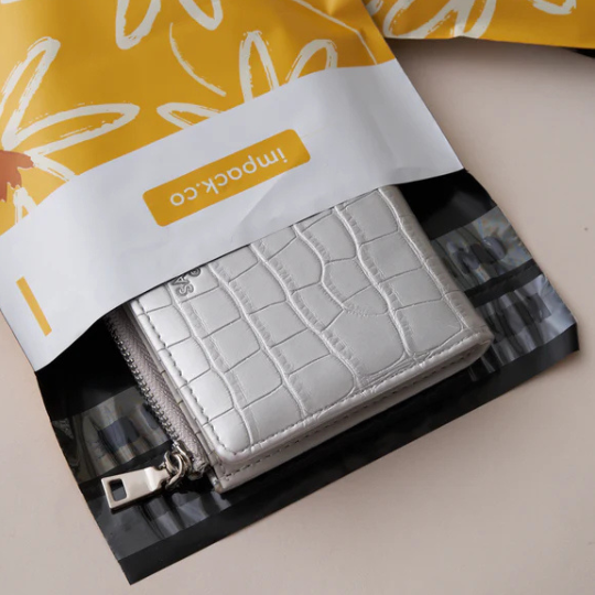 A small silver wallet with a crocodile pattern partially inside a yellow and white floral plastic shipping bag.