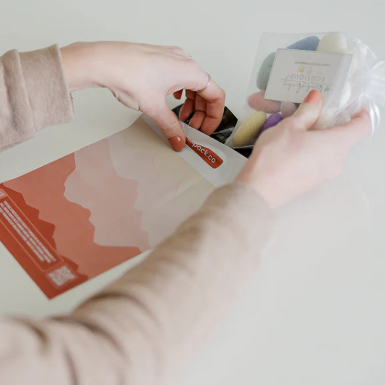 A person’s hands place a small mesh bag with colorful objects and a card into a peach-colored envelope on a white surface.