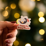 A hand with red painted nails holds an impack.co Christmas Thank You Sticker featuring a penguin in a Santa hat and scarf with “Thank You Snow Much!”—festive for holiday packaging, set before blurred yellow lights.