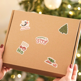A person holds a plain brown box decorated with impack.co Christmas Thank You Stickers in front of a blurred Christmas tree.