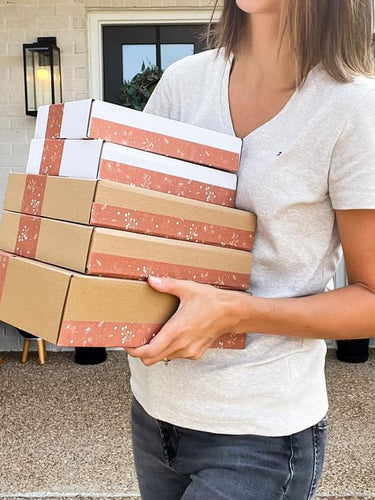 A person in a light gray T-shirt holds a stack of four brown and two white boxes sealed with pink floral tape, standing outside a house with a white brick exterior.