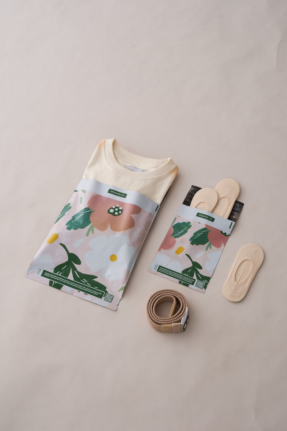 A folded shirt, a folded pair of socks, a rolled-up belt, and a pair of shoe insoles are arranged on a light-colored surface with impack.co Pastel Petals Mailers 10" x 13" nearby for transit protection.