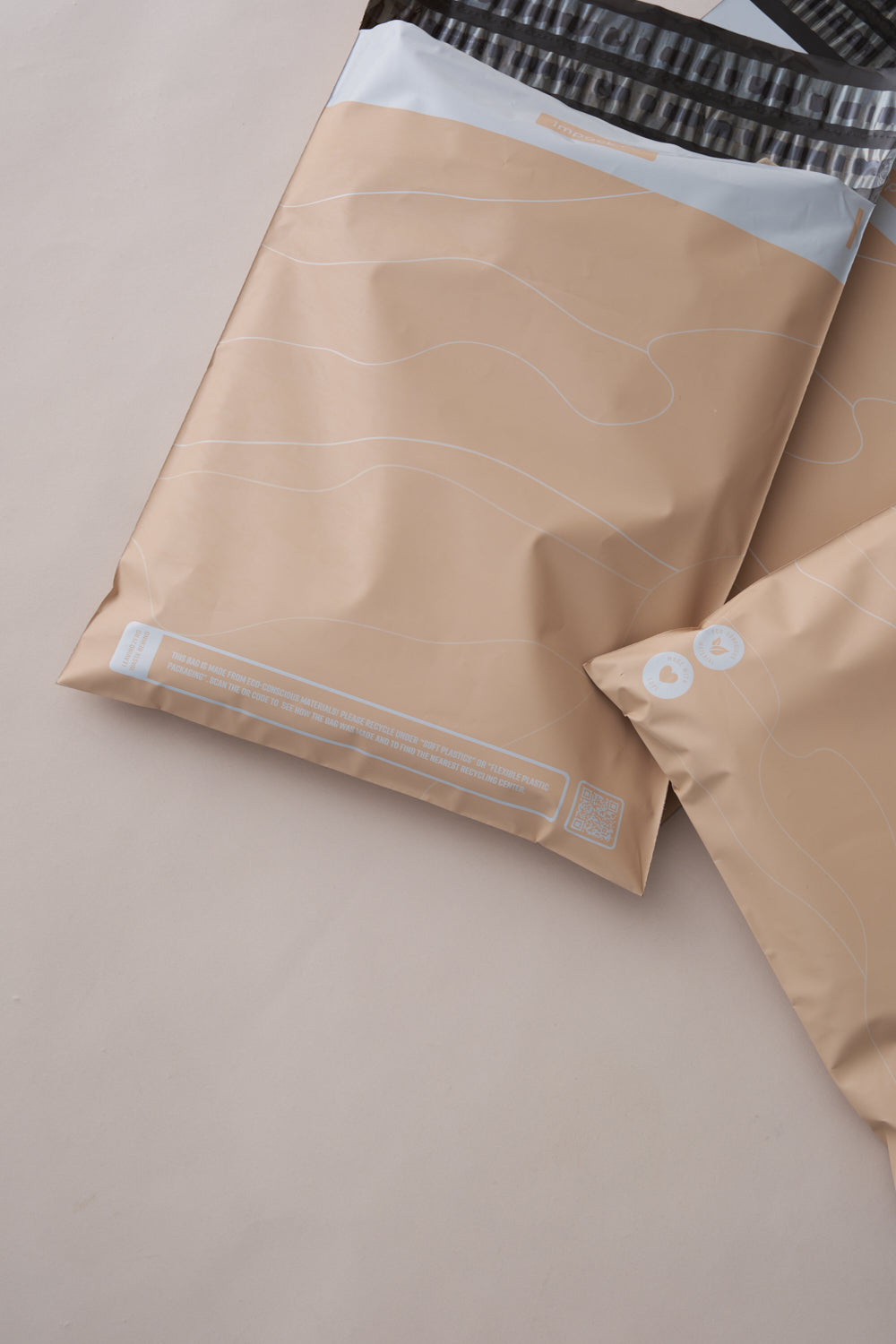 Two tan-colored plastic bags with white patterns and text, placed on a beige surface. One bag, offering versatile packaging solutions, is larger and partially overlaps the smaller one. These are Tidal Sand Mailers 10" x 13" by impack.co.