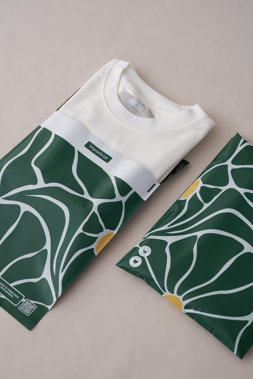 A white T-shirt neatly folded inside green packaging with a white and yellow floral design, ensuring transit protection. Another similar versatile Forest Grove Mailers 10" x 13" by impack.co is next to it.