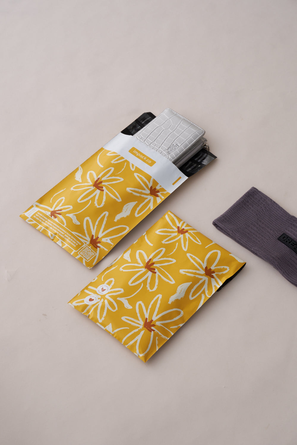 Two impack.co Golden Pencil Floral Mailers 6" x 9" containing items, one partly open showing a silver textured item inside. A gray rectangular cloth is placed to the right of the mailers, ensuring transit protection.