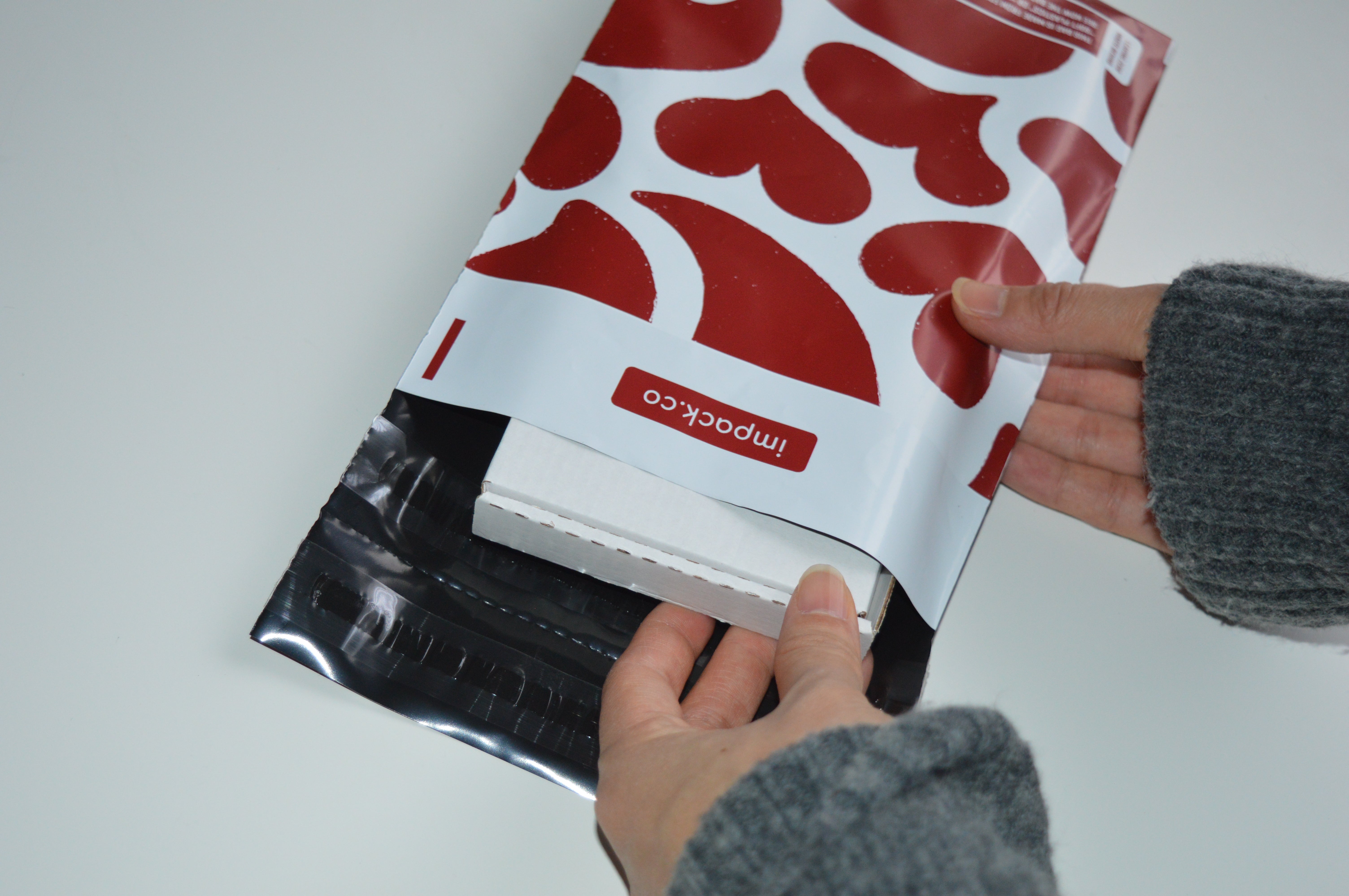 Hands placing a white box into an impack.co Red Hearts Biodegradable Mailer 6" x 9", perfect for jewelry packaging or small shipping envelopes.