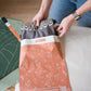 A woman is opening a Floral 2D Rosy Brown Biodegradable Mailers 10" x 13" bag of clothes on a bed.