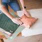 A woman cutting a piece of fabric on a table, using recyclable materials or zero waste techniques to create Olive Leaf Biodegradable Mailers 10" x 13" by impack.co.