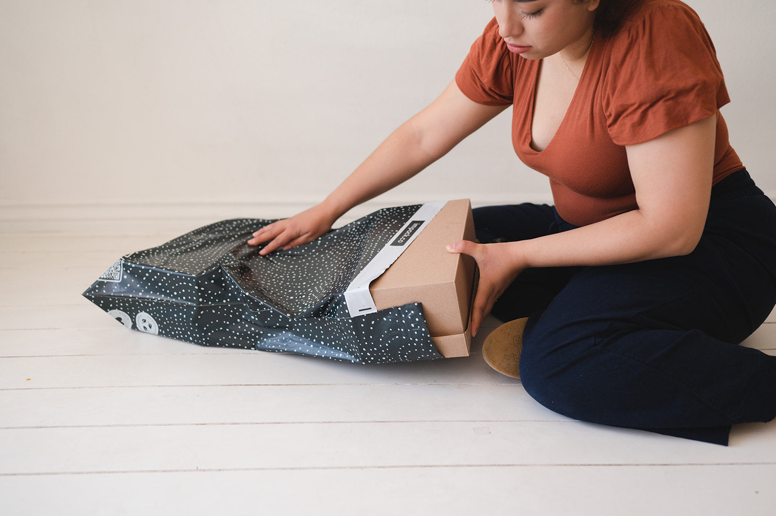 A woman is opening a Wavy Dots Biodegradable Mailers 19" x 24" box from impack.co on the floor.