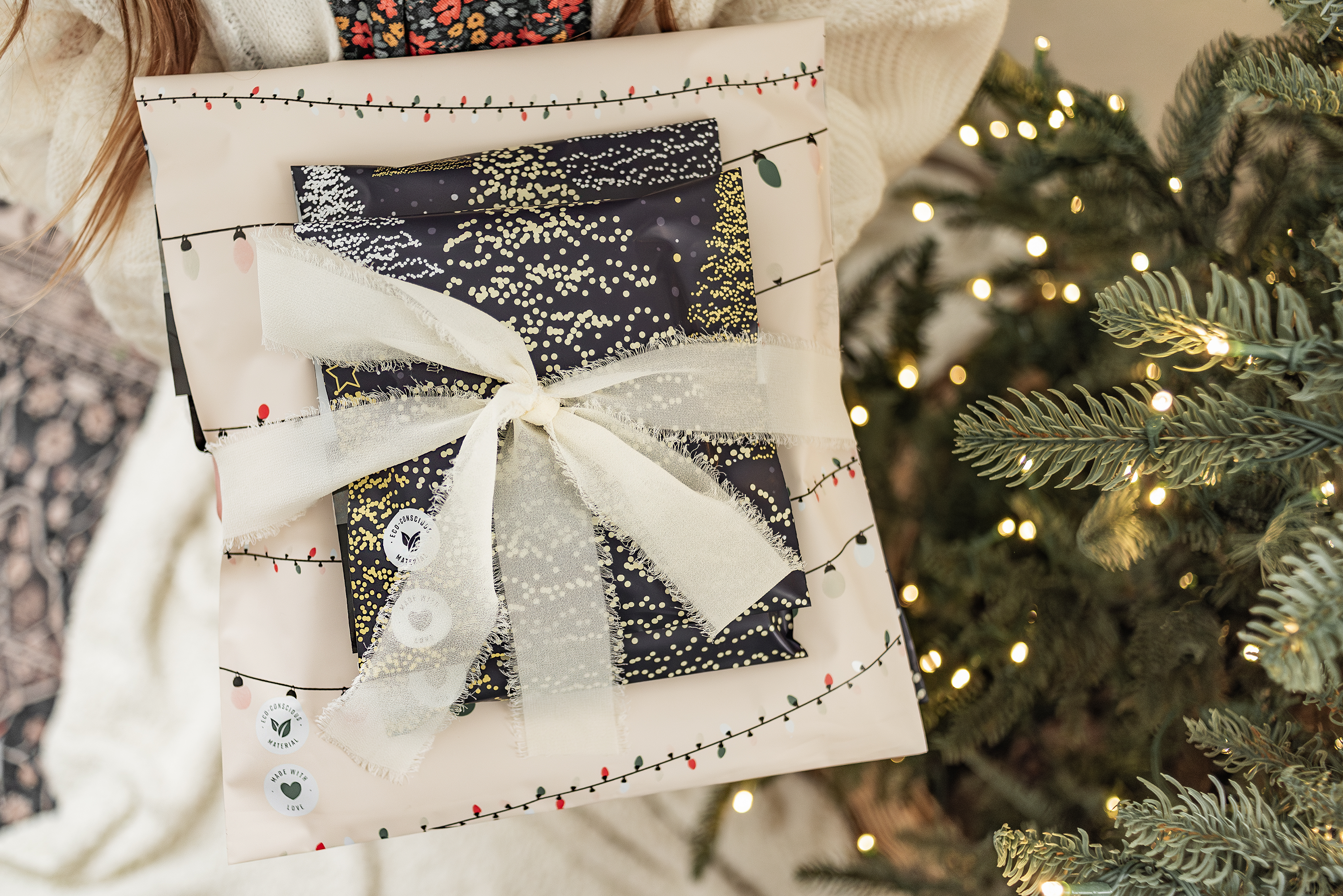 A person holding an impack.co Christmas Tree Biodegradable Mailer, featuring a festive holiday-themed design and a shiny bow, stands next to a Christmas tree adorned with lights. This mailer ensures safe transit protection, keeping the gift secure and ready for joyful unwrapping.