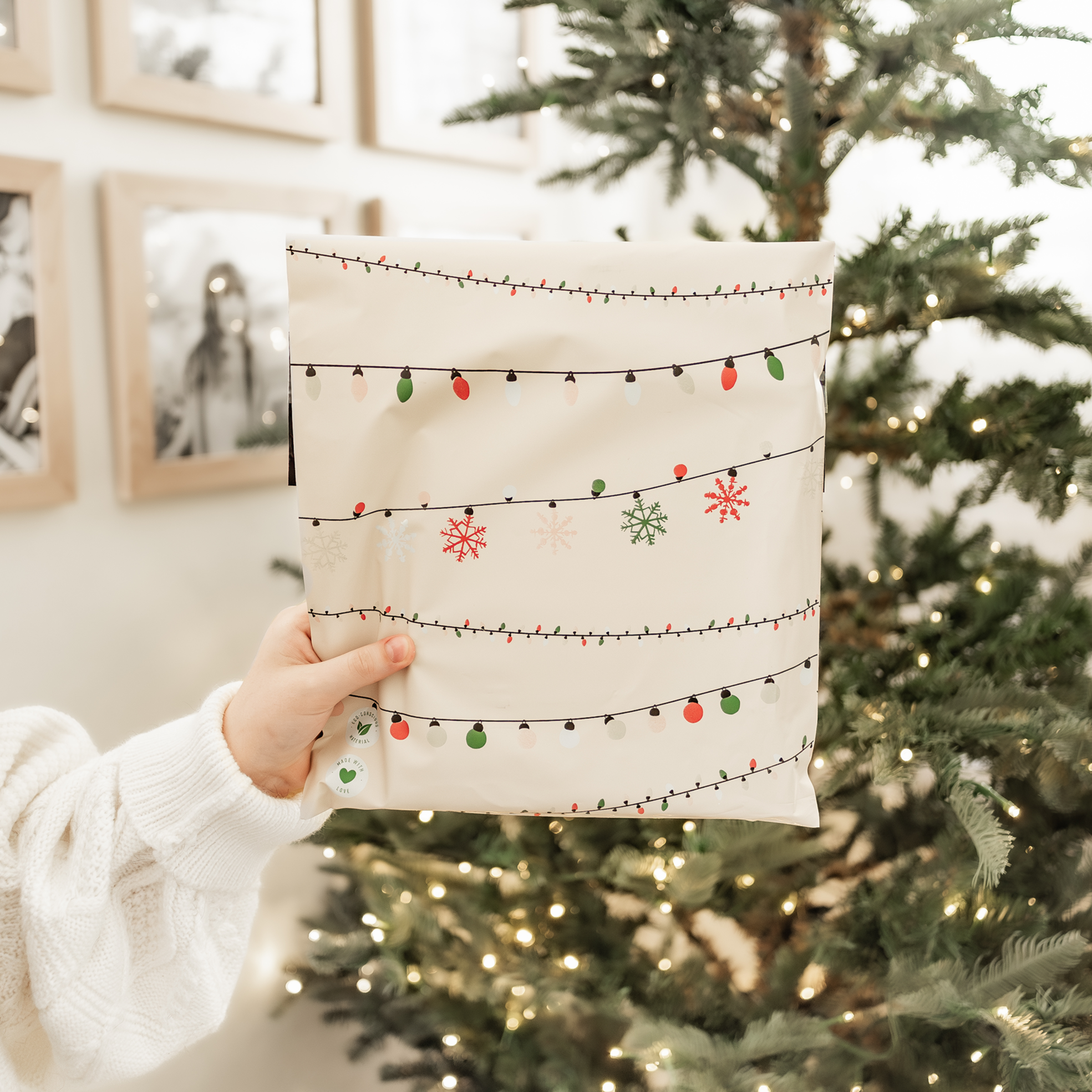 An eco-friendly woman holding up a Christmas Light Strings Biodegradable Mailers 10" x 13" gift bag from impack.co in front of a festive Christmas tree.