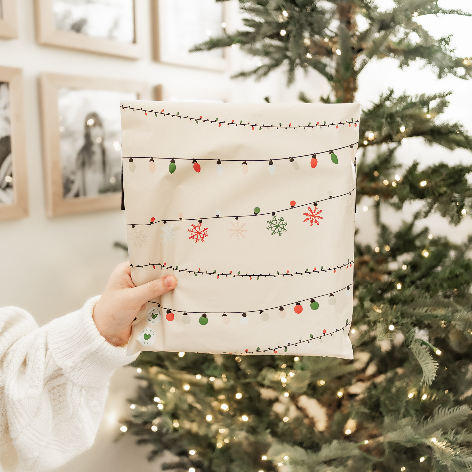 A woman holding up a Christmas Light Strings Biodegradable Mailers 14.5" x 19" eco-friendly gift bag from impack.co in front of a US stock christmas tree.