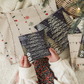 A child is holding a Christmas card on a blanket next to a Christmas tree. The card features vibrant illustrations of Impack.co Christmas Tree Biodegradable Mailers 6" x 9", making it an eco-friendly choice for holiday greetings. Plus