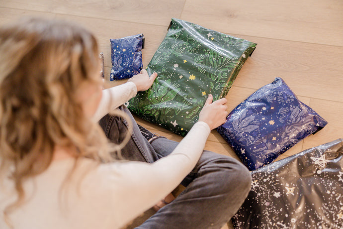 A person sitting on the floor wraps gifts in star-patterned paper, with one green and three blue packages visible.
