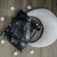 A Midnight Galaxy Mailers 14.5" x 19" pillow with stars and a crescent laying on a wooden floor, made by impack.co.