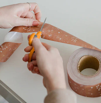 A person cuts a strip of brown floral-patterned tape with scissors near a white table; a roll of the same tape sits beside them.