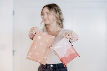 A person holds two impack.co envelopes, one with Celestial Tan moon and star prints and the other featuring a wave design, set against a blurred background.