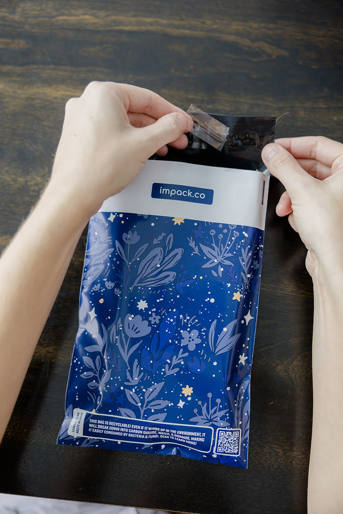 Hands opening a package with a blue floral pattern labeled "impack.co" on a dark wooden table, featuring the SALE - Midnight Indigo Mailers 6" x 9", offering durable and sustainable biodegradable protection.