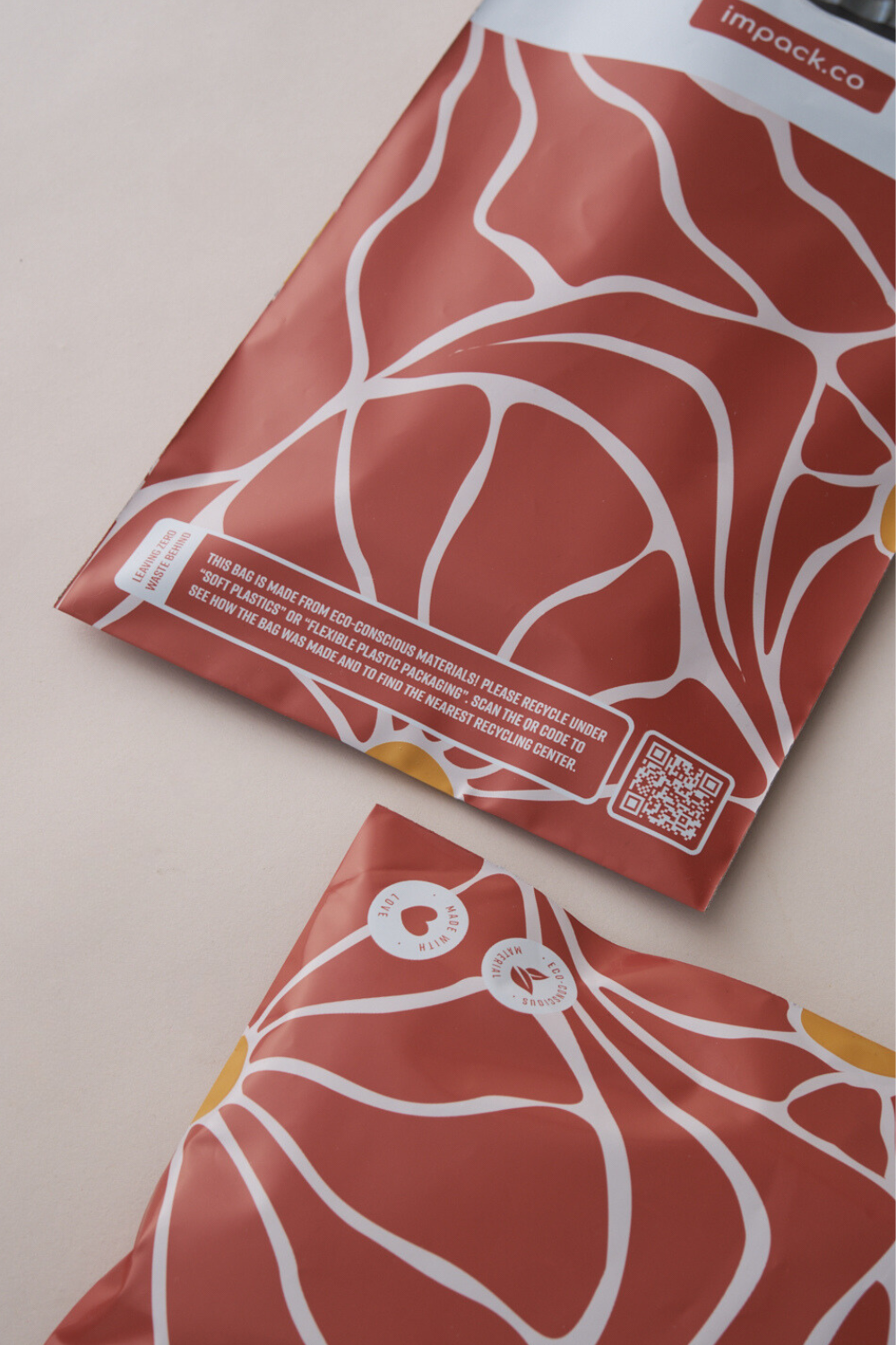 Two red packaging bags with a white abstract design, featuring the Crimson Grove print, are placed on a light surface. One bag is partially shown. Both have labels and QR codes, showcasing these Crimson Crove Mailers 10" x 13" by impack.co as a versatile packaging solution.