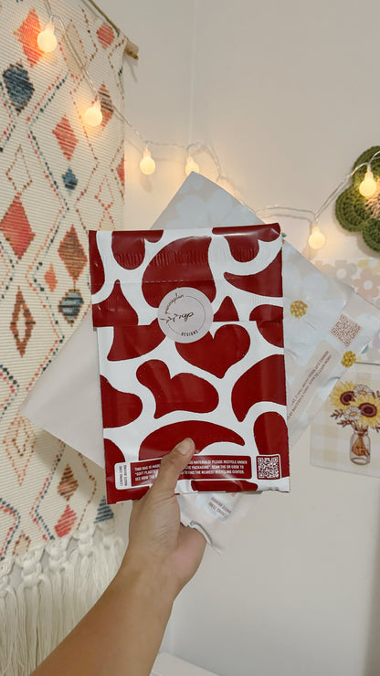 A hand holding a decorative packaging envelope of impack.co's Red Hearts Biodegradable Mailers 10" x 13" against a bohemian-inspired interior background with string lights.