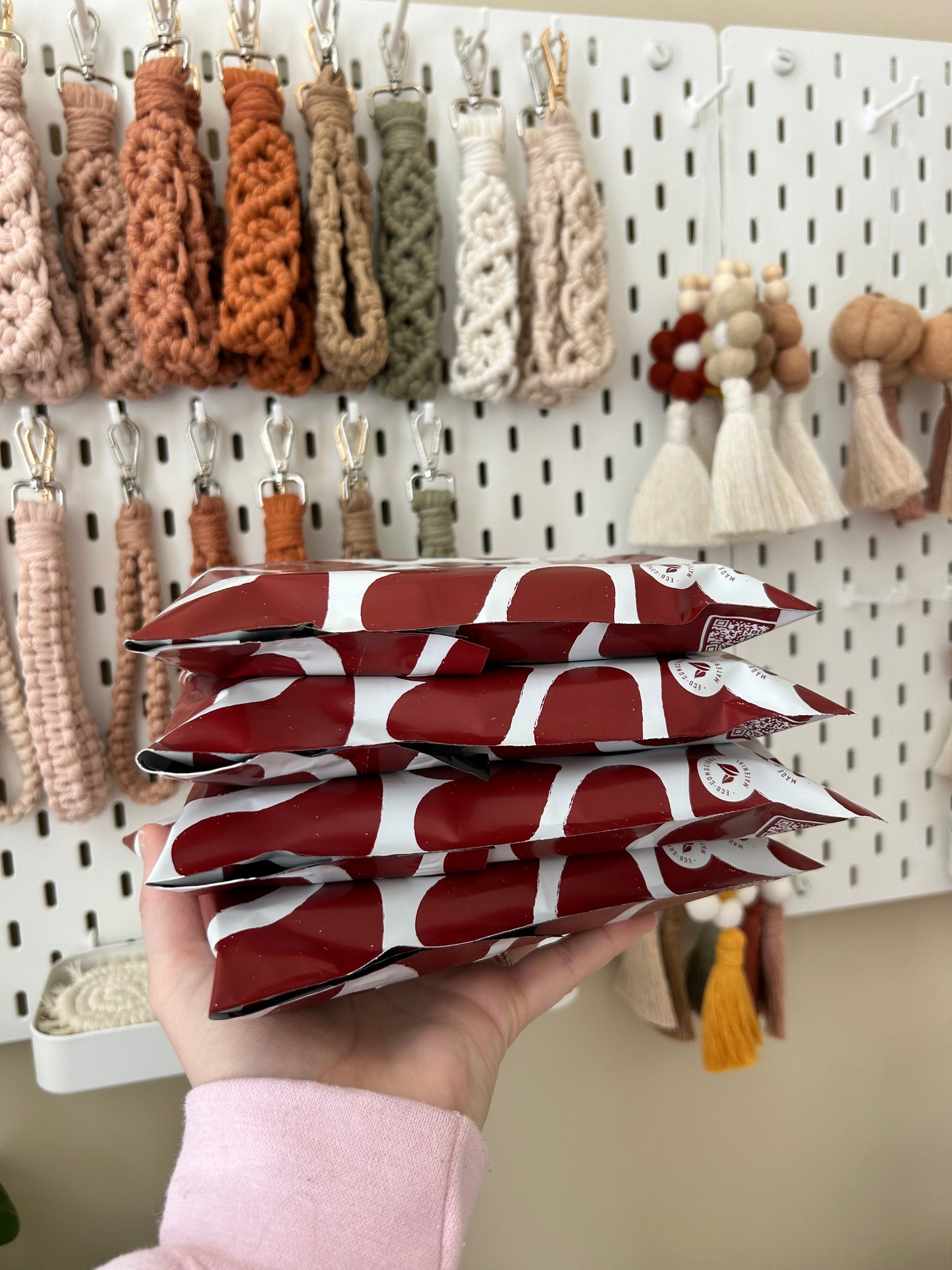 A person holding a stack of Impack.co Red Hearts Biodegradable Mailers 10" x 13" in front of a wall display with a variety of tasseled keychains.