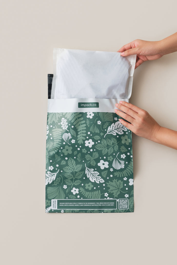 Hands placing a Glassine Bags 9.6" x 12.6" into a green and white floral-patterned shipping bag labeled "impack.co".