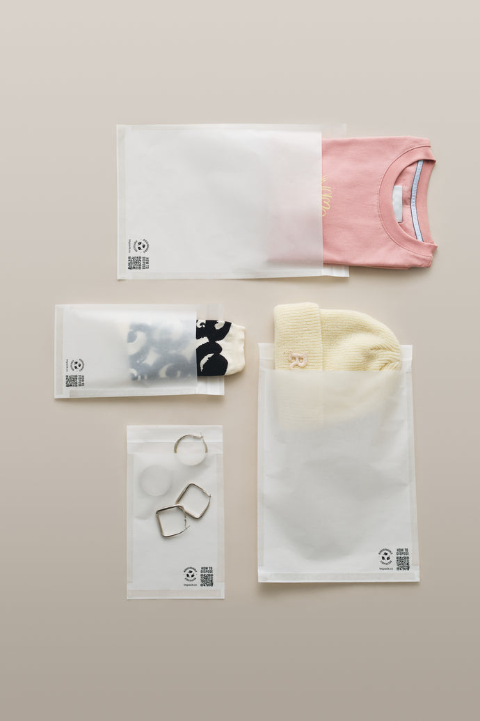 White and pink T-shirt, yellow beanie, black and white scarf, silver rings, and two geometric earrings are arranged on a light beige surface, each item partially inside translucent impack.co Glassine Bags 9.6" x 12.6".