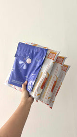 A hand holds four impack.co mailers—three are white with orange accents, while one is a Boho Sun & Moon Biodegradable Mailer (14.5" x 19"), perfect for shipping bulkier items sustainably.