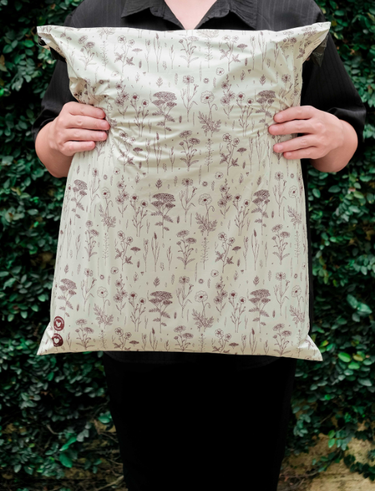 A person in dark clothing holds a large, sealed package with a floral pattern design, standing in front of leafy green foliage.