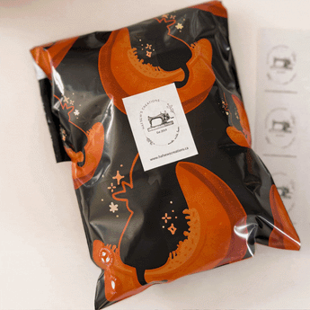 A sealed black and orange package with illustrations and a sticker label featuring a sewing machine logo and website address. A sheet of similar stickers lies nearby.