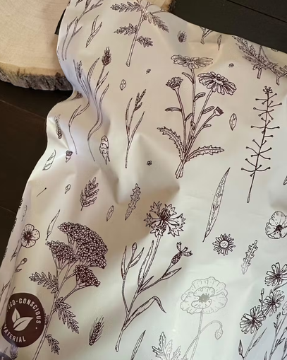 A crumpled mailer with illustrated botanical designs rests on a dark surface next to a round piece of wood. An "eco-conscious material" label is visible on the mailer.