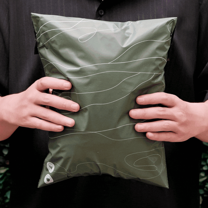 A person in a dark shirt holds a large, sealed green package with a white abstract design, gripping it with both hands.