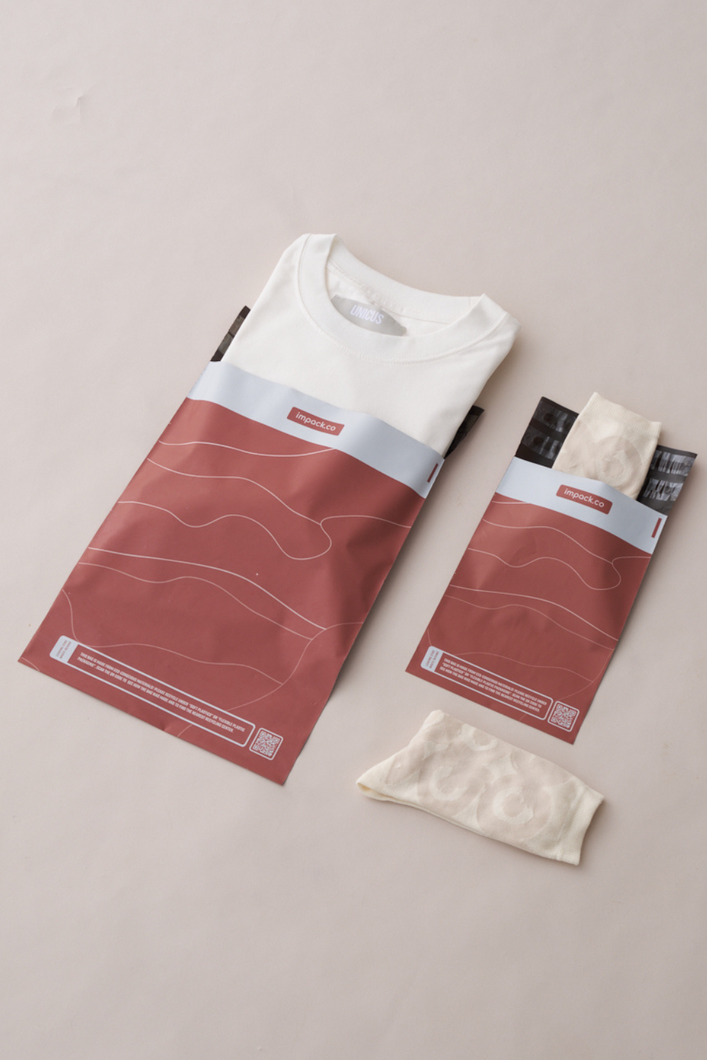 Folded white t-shirt in a brown paper bag, accompanied by a pair of beige socks beside their matching packaging, all placed on a light neutral surface—perfectly suited for medium-sized shipments using impack.co Tidal Terracotta Mailers 10" x 13".