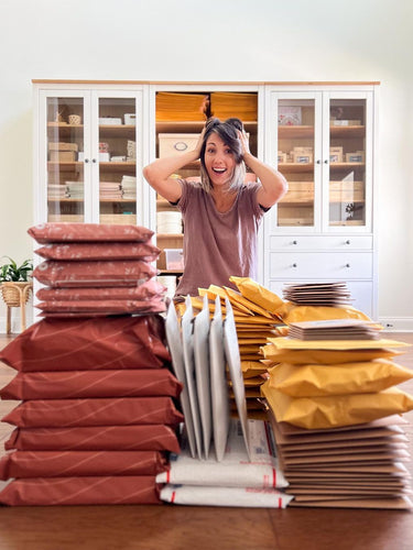 Person excitedly standing behind stacks of variously colored and sized packages on a wooden floor in front of a white cabinet.
