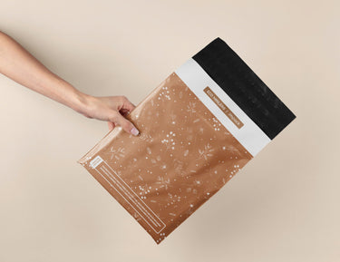A hand holds impack.co’s SALE - Floral 2D Rosy Brown Biodegradable Mailer (10" x 13") with a black adhesive strip, highlighting eco-friendly, stylish packaging ideal for secure shipping on a beige background.