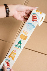 A hand holds an impack.co Sticker Roll with positive phrases like "Thank you" and "Cool thing inside," above stacked boxes. These eco-friendly rolls add a cheerful touch to every package.