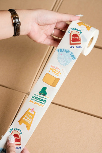 A hand holds an impack.co Sticker Roll with positive phrases like "Thank you" and "Cool thing inside," above stacked boxes. These eco-friendly rolls add a cheerful touch to every package.