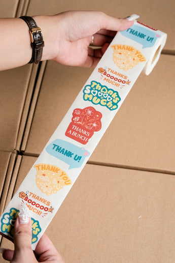 A person holds an impack.co Sticker Roll featuring assorted colorful "Thank You" packaging stickers above a cardboard surface.
