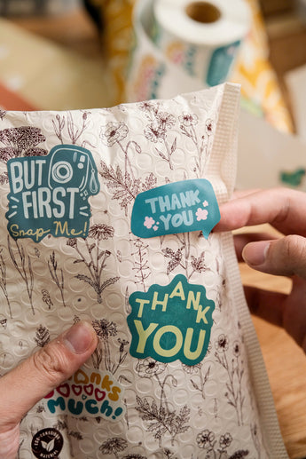 A person holds an impack.co Sticker Roll package with floral designs, decorated with recyclable paper stickers that say "THANK YOU," "BUT FIRST Snap Me!," and "thanks so much!" A roll of stickers is visible in the background.