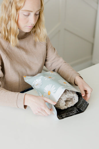 A person sits at a table, holding an impack.co Daisy White Biodegradable Mailer (10" x 13"), which contains a skein of yarn.