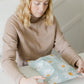 A woman is sitting at a table with a Daisy White Biodegradable Mailers 10" x 13" bag from impack.co in front of her.