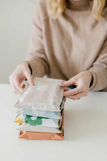 A person in a beige sweater is arranging a stack of colorful Cosmos Biodegradable Mailers by impack.co on a white surface.