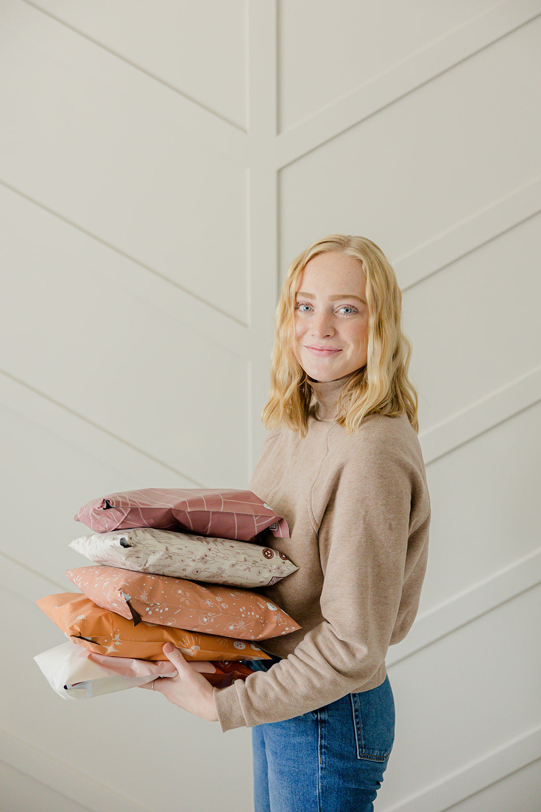 A woman holding a stack of mailer bags 6" x 9" pillows in front of a white wall.