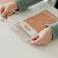 A woman is holding a Rosy Brown Biodegradable Bubble Mailer 6" x 9" from impack.co on a table.